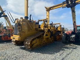 A lineup of heavy construction machinery, including large bulldozers and other vehicles, set on a gravel surface. The equipment features prominent treads and metal arms, indicating their use for construction and heavy-duty tasks.