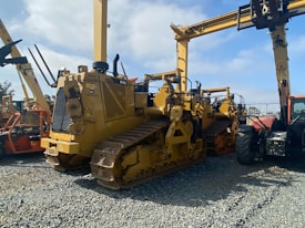 A lineup of heavy construction machinery, including large bulldozers and other vehicles, set on a gravel surface. The equipment features prominent treads and metal arms, indicating their use for construction and heavy-duty tasks.