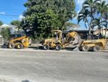 A construction scene with heavy machinery positioned on a street lined with large green trees and bushes. The yellow machines include a skid steer loader and a trencher parked on the road. In the background, there is a house partially obscured by the trees.