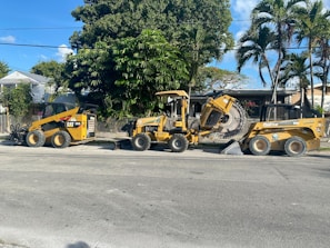 A construction scene with heavy machinery positioned on a street lined with large green trees and bushes. The yellow machines include a skid steer loader and a trencher parked on the road. In the background, there is a house partially obscured by the trees.