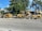 A construction scene with heavy machinery positioned on a street lined with large green trees and bushes. The yellow machines include a skid steer loader and a trencher parked on the road. In the background, there is a house partially obscured by the trees.