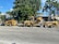 A construction scene with heavy machinery positioned on a street lined with large green trees and bushes. The yellow machines include a skid steer loader and a trencher parked on the road. In the background, there is a house partially obscured by the trees.