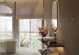 A modern, elegant bathroom with a clean white bathtub situated near a large window that lets in natural light. A decorative bouquet of vibrant red and pink flowers sits on a dark wooden shelf, accompanied by a sleek round washbasin and a stylish faucet. The wall features a large, oval mirror with backlighting, and two hooks are mounted beside it. The overall ambiance is serene and inviting.