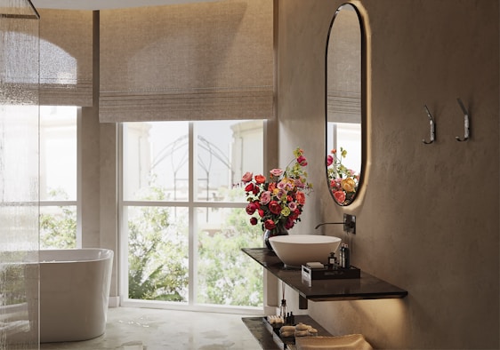 A modern, elegant bathroom with a clean white bathtub situated near a large window that lets in natural light. A decorative bouquet of vibrant red and pink flowers sits on a dark wooden shelf, accompanied by a sleek round washbasin and a stylish faucet. The wall features a large, oval mirror with backlighting, and two hooks are mounted beside it. The overall ambiance is serene and inviting.