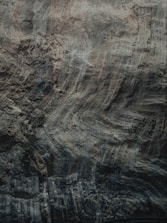 The image features a close-up view of a rock surface, highlighting its natural textures and striations. The surface displays a variety of swirling patterns and layered ridges, giving it an intricate and rugged appearance. The colors are muted and earthy, with shades of gray, brown, and hints of green.