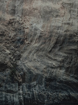The image features a close-up view of a rock surface, highlighting its natural textures and striations. The surface displays a variety of swirling patterns and layered ridges, giving it an intricate and rugged appearance. The colors are muted and earthy, with shades of gray, brown, and hints of green.
