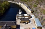 Aerial view of an industrial facility located alongside a narrow body of water. The facility consists of several long, yellow buildings with grey roofs, surrounded by hills with sparse vegetation. The water has a deep blue color, bordered by a mix of rocky terrain and a small, wooded area.