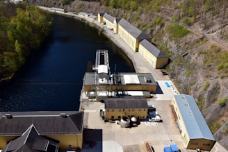 Aerial view of an industrial facility located alongside a narrow body of water. The facility consists of several long, yellow buildings with grey roofs, surrounded by hills with sparse vegetation. The water has a deep blue color, bordered by a mix of rocky terrain and a small, wooded area.