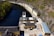 Aerial view of an industrial facility located alongside a narrow body of water. The facility consists of several long, yellow buildings with grey roofs, surrounded by hills with sparse vegetation. The water has a deep blue color, bordered by a mix of rocky terrain and a small, wooded area.