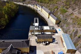 Aerial view of an industrial facility located alongside a narrow body of water. The facility consists of several long, yellow buildings with grey roofs, surrounded by hills with sparse vegetation. The water has a deep blue color, bordered by a mix of rocky terrain and a small, wooded area.