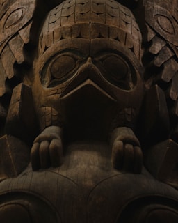 A detailed wooden carving featuring a stylized animal figure, possibly a bird, with intricate patterns and textures. The carving includes prominent features like large eyes, a beak, and detailed feather-like engravings. The wood's natural grain and color add depth to the piece.