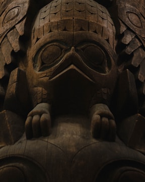 A detailed wooden carving featuring a stylized animal figure, possibly a bird, with intricate patterns and textures. The carving includes prominent features like large eyes, a beak, and detailed feather-like engravings. The wood's natural grain and color add depth to the piece.