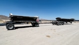 A large dump trailer with multiple wheels sits on a sandy desert landscape under a clear blue sky. Mountains are visible in the background, and the land is barren with sparse vegetation.