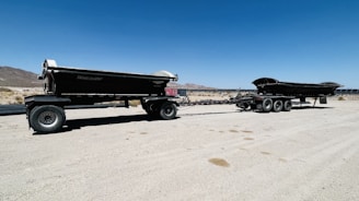 A large dump trailer with multiple wheels sits on a sandy desert landscape under a clear blue sky. Mountains are visible in the background, and the land is barren with sparse vegetation.