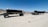 A large dump trailer with multiple wheels sits on a sandy desert landscape under a clear blue sky. Mountains are visible in the background, and the land is barren with sparse vegetation.