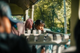 A person is sitting inside a tram or bus, gazing thoughtfully out the window. The light streaming through casts shadows and highlights, creating a warm and serene atmosphere. The background shows blurred greenery outside, suggesting movement and travel.