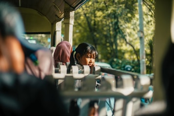 A person is sitting inside a tram or bus, gazing thoughtfully out the window. The light streaming through casts shadows and highlights, creating a warm and serene atmosphere. The background shows blurred greenery outside, suggesting movement and travel.