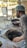 A man wearing a hair net is making flatbreads in a traditional tandoor oven. The scene is set outdoors with metal tables nearby. The background shows a street with a sidewalk and some greenery.