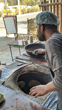 A man wearing a hair net is making flatbreads in a traditional tandoor oven. The scene is set outdoors with metal tables nearby. The background shows a street with a sidewalk and some greenery.