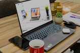 A laptop with its screen on displaying a desktop wallpaper featuring multiple devices is placed on a wooden table. Surrounding the laptop are various items, including a smartphone, a red coffee cup, a mouse, a notebook with pens, a small potted succulent plant, earphones, and a few decorative candles.