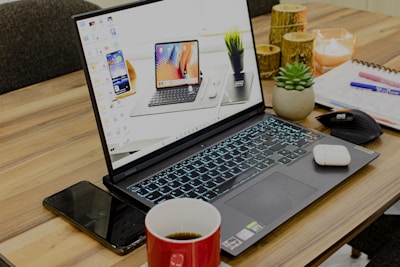 A laptop with its screen on displaying a desktop wallpaper featuring multiple devices is placed on a wooden table. Surrounding the laptop are various items, including a smartphone, a red coffee cup, a mouse, a notebook with pens, a small potted succulent plant, earphones, and a few decorative candles.