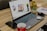 A laptop with its screen on displaying a desktop wallpaper featuring multiple devices is placed on a wooden table. Surrounding the laptop are various items, including a smartphone, a red coffee cup, a mouse, a notebook with pens, a small potted succulent plant, earphones, and a few decorative candles.