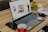 A laptop with its screen on displaying a desktop wallpaper featuring multiple devices is placed on a wooden table. Surrounding the laptop are various items, including a smartphone, a red coffee cup, a mouse, a notebook with pens, a small potted succulent plant, earphones, and a few decorative candles.