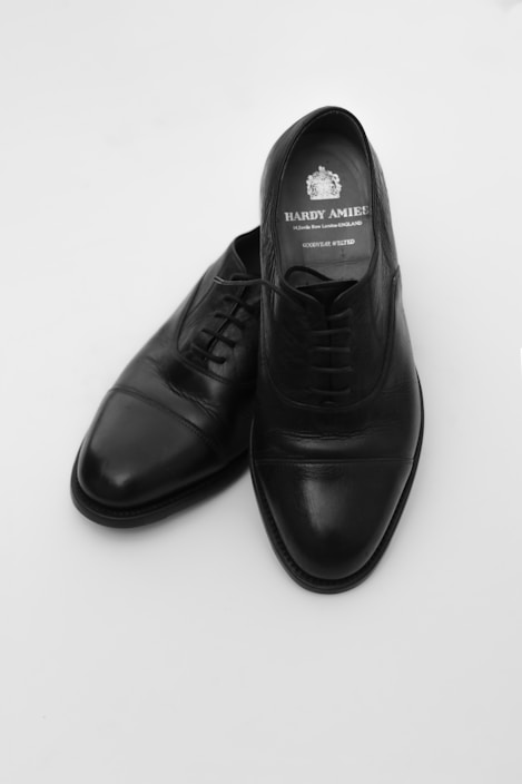 A pair of black leather dress shoes placed against a plain white background. The shoes are elegant with a polished finish and a classic design, featuring laces and a slightly pointed toe. The insole displays a brand logo and name.