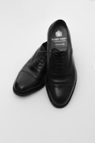 A pair of black leather dress shoes placed against a plain white background. The shoes are elegant with a polished finish and a classic design, featuring laces and a slightly pointed toe. The insole displays a brand logo and name.