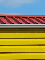 A close-up view of a building section with a corrugated red metal roof and bright yellow horizontal siding panels against a blue sky.