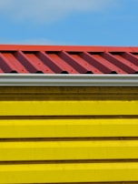 A close-up view of a building section with a corrugated red metal roof and bright yellow horizontal siding panels against a blue sky.