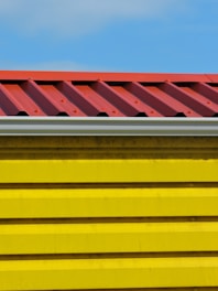 A close-up view of a building section with a corrugated red metal roof and bright yellow horizontal siding panels against a blue sky.