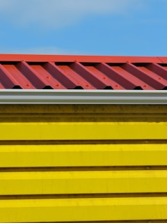 A close-up view of a building section with a corrugated red metal roof and bright yellow horizontal siding panels against a blue sky.