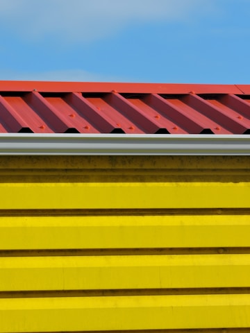 A close-up view of a building section with a corrugated red metal roof and bright yellow horizontal siding panels against a blue sky.