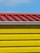 A close-up view of a building section with a corrugated red metal roof and bright yellow horizontal siding panels against a blue sky.