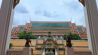 A traditional Thai temple roof with ornate, tiered green and red sections, bordered by decorative elements. In the foreground, stone sculptures and intricate architectural designs, including a small pagoda and guardian statues, frame the view.