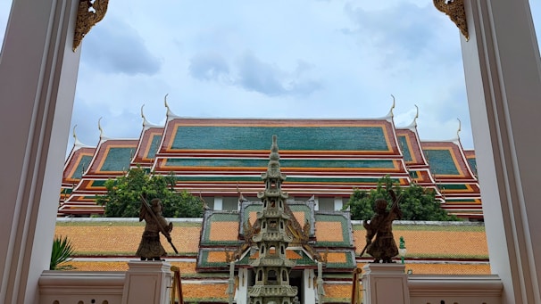 A traditional Thai temple roof with ornate, tiered green and red sections, bordered by decorative elements. In the foreground, stone sculptures and intricate architectural designs, including a small pagoda and guardian statues, frame the view.