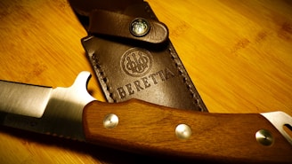 A close-up view of a finely crafted knife with a wooden handle lying beside a leather sheath, which has the brand name and logo embossed on it. The blade appears sharp and well-polished, and the leather is stitched with precision.