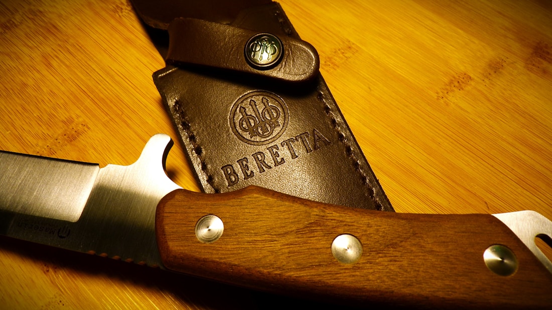 A close-up view of a finely crafted knife with a wooden handle lying beside a leather sheath, which has the brand name and logo embossed on it. The blade appears sharp and well-polished, and the leather is stitched with precision.