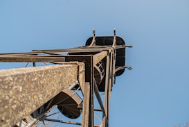 A low-angle view of a metal tower structure with a clear blue sky in the background. The tower has several beams and a spiral staircase winding up through its center.