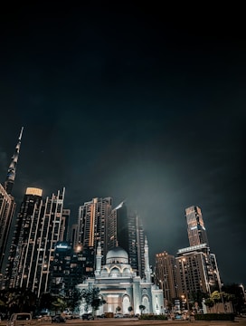 A night view of a cityscape with modern skyscrapers illuminated by lights. In the foreground, a large mosque with ornate architecture stands out with its bright illumination. The sky is dark, accentuating the glow from the buildings.