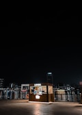 A small, illuminated food kiosk named 'Lokma' is situated near a waterfront at night. The kiosk is lit up and stands out against the dark sky and water. In the background, city lights reflect on the water, and tall buildings are visible, giving a sense of an urban environment.