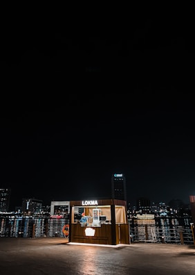 A small, illuminated food kiosk named 'Lokma' is situated near a waterfront at night. The kiosk is lit up and stands out against the dark sky and water. In the background, city lights reflect on the water, and tall buildings are visible, giving a sense of an urban environment.