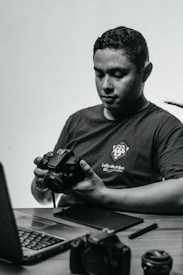 A person sits at a desk holding a camera, looking down at it in concentration. On the desk are a laptop and various camera equipment and accessories, including lenses and a pen tablet.
