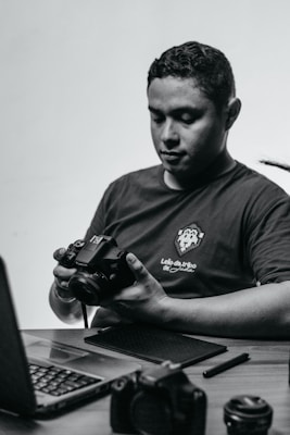 A person sits at a desk holding a camera, looking down at it in concentration. On the desk are a laptop and various camera equipment and accessories, including lenses and a pen tablet.