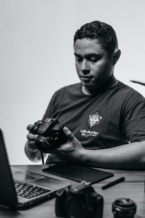 A person sits at a desk holding a camera, looking down at it in concentration. On the desk are a laptop and various camera equipment and accessories, including lenses and a pen tablet.