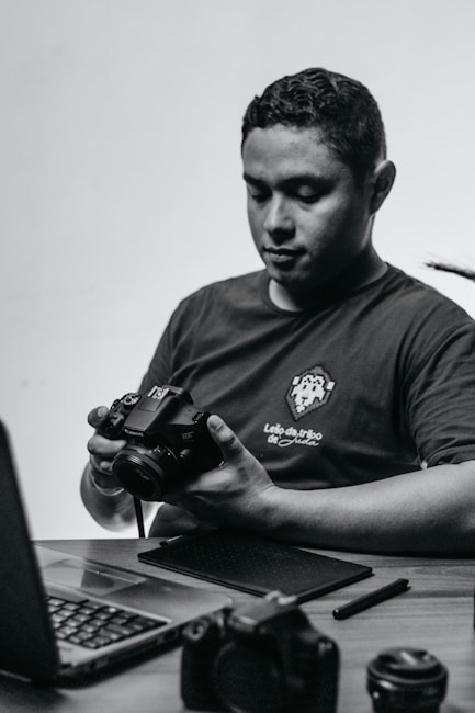 A person sits at a desk holding a camera, looking down at it in concentration. On the desk are a laptop and various camera equipment and accessories, including lenses and a pen tablet.