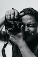 A person holds a camera close to their face with one eye closed, concentrating on taking a photo. The image is in black and white, highlighting the contrast and texture on the person's face and hands.