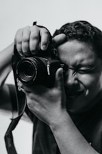 A person holds a camera close to their face with one eye closed, concentrating on taking a photo. The image is in black and white, highlighting the contrast and texture on the person's face and hands.