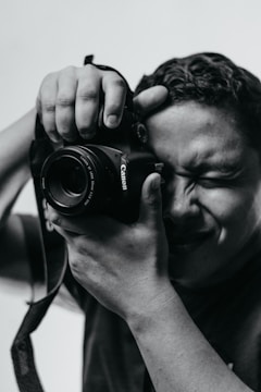A person holds a camera close to their face with one eye closed, concentrating on taking a photo. The image is in black and white, highlighting the contrast and texture on the person's face and hands.